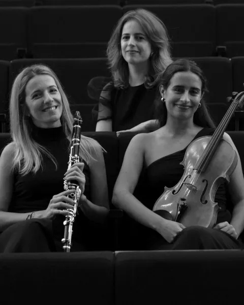 Fotografia a preto e branco das três músicas do Re:Flexus Trio sentadas numa plateia, uma com um clarinete e outra com uma viola d'arco.