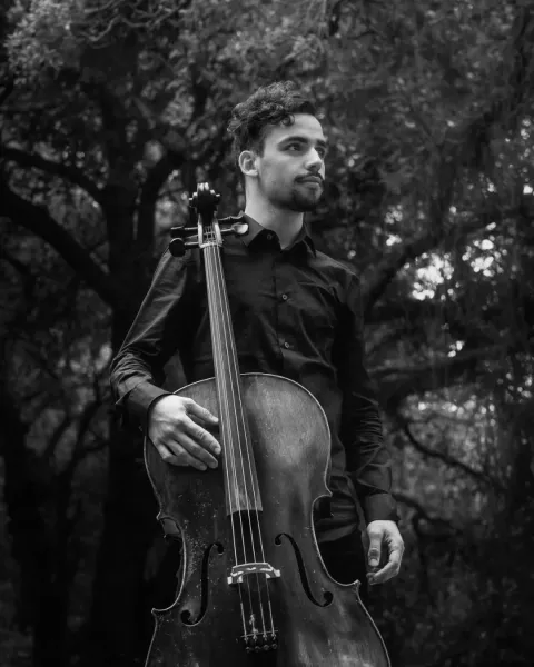 Fotografia a preto e branco de Tiago Azevedo e Silva, de pé entre árvores, a segurar um violoncelo.
