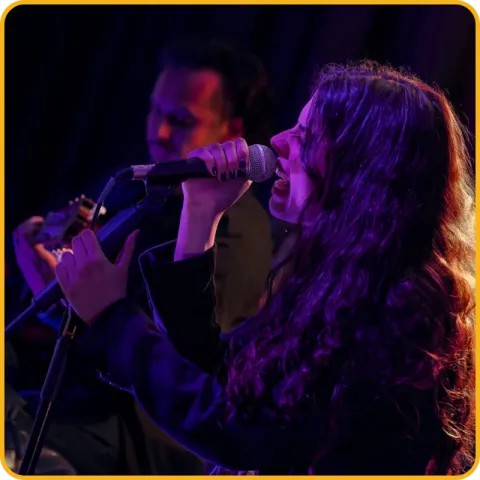 Uma cantora a cantar ao microfone num palco com luz roxa.