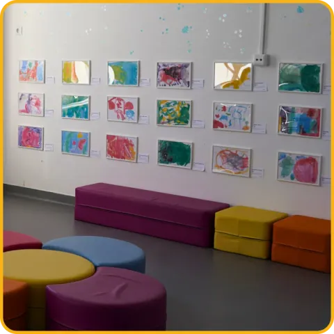 Desenhos coloridos de crianças pendurados numa parede branca, com pufes coloridos em frente.
