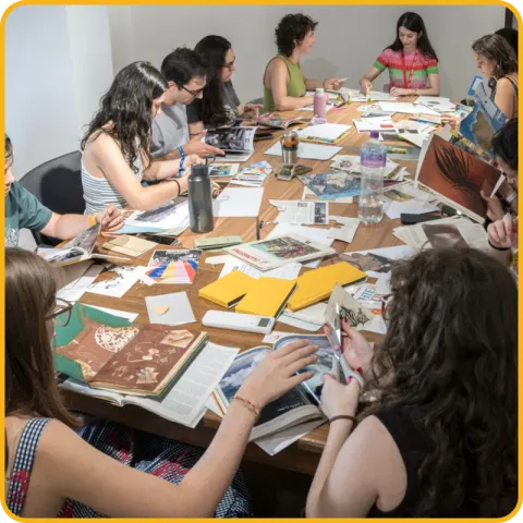 Jovens à volta de uma mesa comprida, a recortar livros e revistas para fazer colagens.