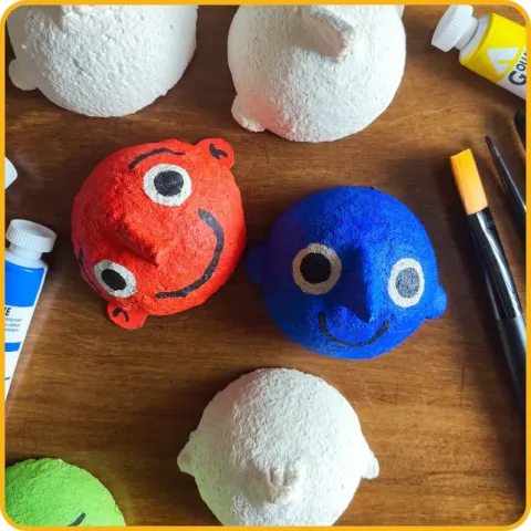 Cabeças de papel machê numa mesa com tintas: uma pintada de vermelho e outra de azul, com caras sorridentes, e outras ainda brancas.