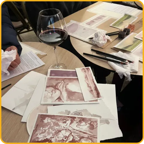 Uma mesa com um copo de vinho tinto, pincéis e pinturas em tons de vinho sobre papel.